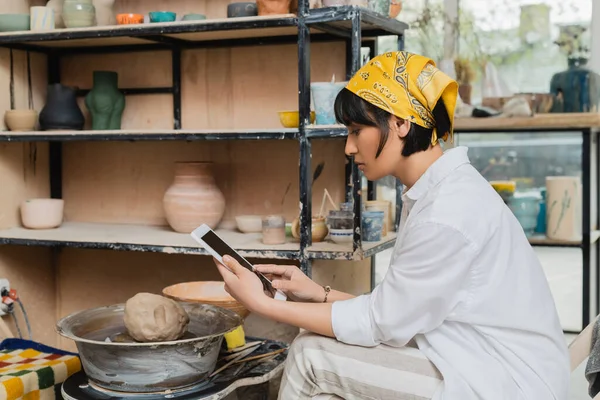 Side view of young brunette asian female artist in headscarf and workwear using digital tablet with blank screen while sitting in ceramic studio at background, craftsmanship in pottery making — Stock Photo