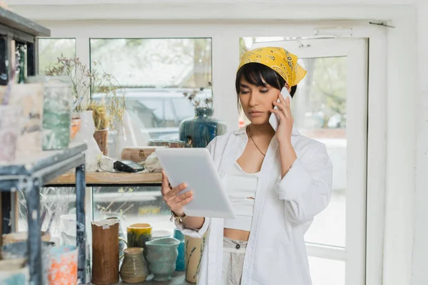 Young asian female artist in headscarf talking on smartphone and using digital tablet while working in blurred ceramic workshop at background, creative process of pottery making — Stock Photo