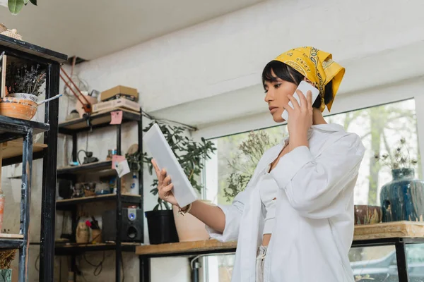 Young brunette asian female potter in headscarf and workwear talking on smartphone and holding digital tablet in blurred ceramic workshop, creative process of pottery making — Stock Photo