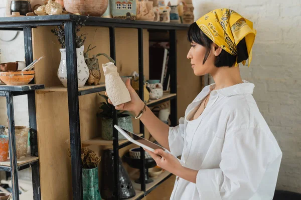 Side view of young asian female artisan in headscarf and workwear holding digital tablet with blank screen and clay sculpture and standing in ceramic workshop, creative process of pottery making — Stock Photo