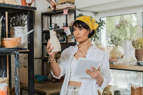 Young brunette asian female artisan in headscarf and workwear holding clay sculpture and digital tablet while working in ceramic workshop, creative process of pottery making — Stock Photo