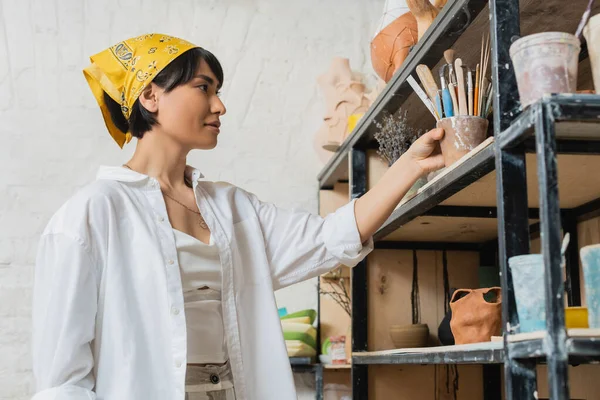 Jovem asiática artista feminina em vestuário de trabalho e lenço de cabeça colocando ferramentas de cerâmica na prateleira perto de produtos de argila e trabalhando em oficina de cerâmica, processo criativo de fabricação de cerâmica — Fotografia de Stock