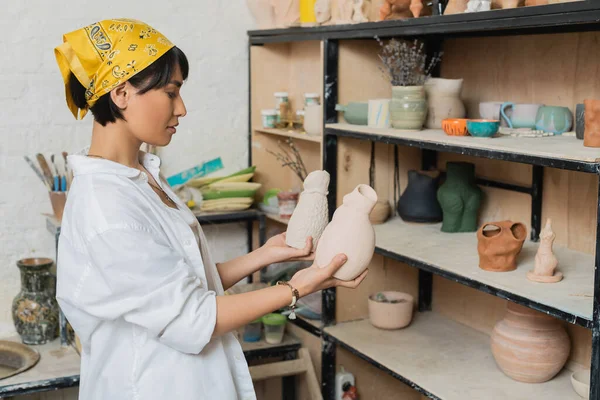 Side view of young asian female potter in headscarf and workwear holding clay sculptures while standing near rack in ceramic workshop, pottery studio scene with skilled artisan — Stock Photo
