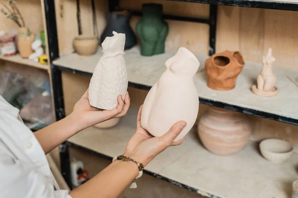 Cropped view of young female artisan in workwear holding clay sculptures while standing near blurred rack in ceramic workshop, pottery studio scene with skilled artisan — Stock Photo