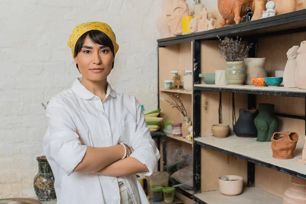 Portrait of young asian female artisan in headscarf and workwear crossing arms and looking at camera near rack with clay sculptures at background, pottery studio scene with skilled artisan — Stock Photo