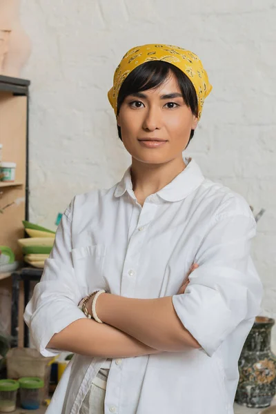 Retrato de jovem asiático oleiro feminino em véu e workwear cruzando braços e olhando para a câmera enquanto em pé na oficina de cerâmica, cerâmica estúdio cena com hábil artesão — Fotografia de Stock
