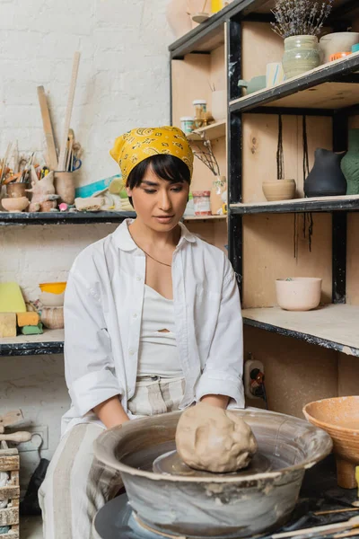 Young asian female artisan in headscarf and workwear looking at clay on pottery wheel while sitting near rack with sculptures in workshop, pottery studio scene with skilled artisan — Stock Photo