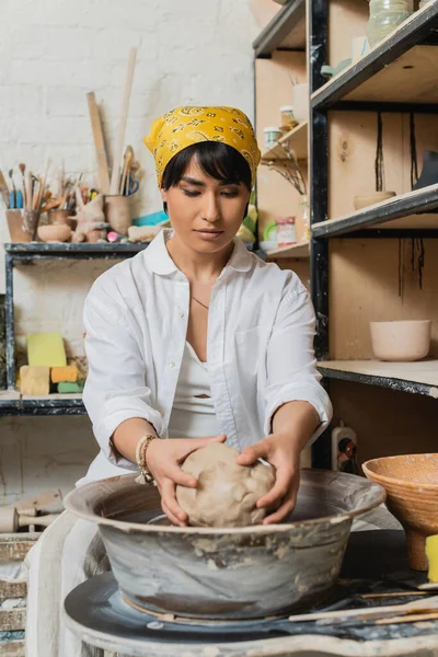 Young brunette asian artisan in headscarf and workwear holding clay while working with pottery wheel near rack and blurred pottery tools in workshop, pottery studio scene with skilled artisan — Stock Photo