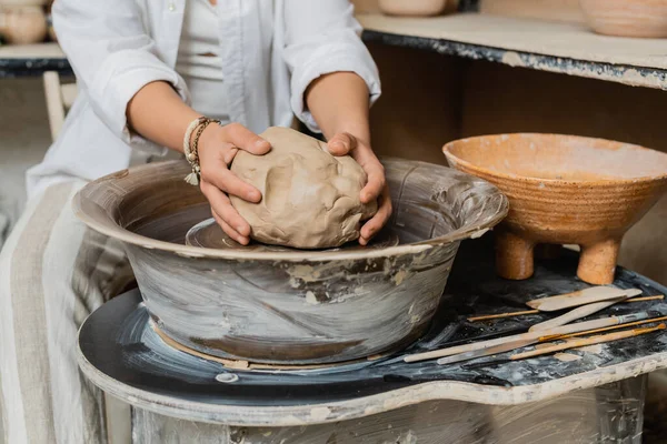 Cropped view of blurred female artisan in workwear putting clay on pottery wheel near tools and bowl in ceramic workshop, pottery studio scene with skilled artisan — Stock Photo
