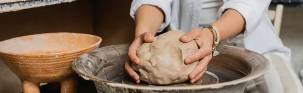 Vue recadrée d'une artiste féminine en vêtements de travail mettant de l'argile sur une roue de poterie près d'un bol tout en travaillant dans un atelier de céramique en arrière-plan, scène d'atelier de poterie avec un artisan qualifié, bannière — Photo de stock