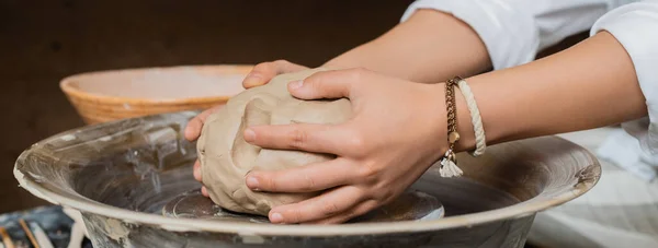 Обрезанный вид молодой женщины гончара положить глину на гончарное колесо во время работы возле чаши в размытой керамической мастерской, ремесленная керамика в студии, баннер — стоковое фото
