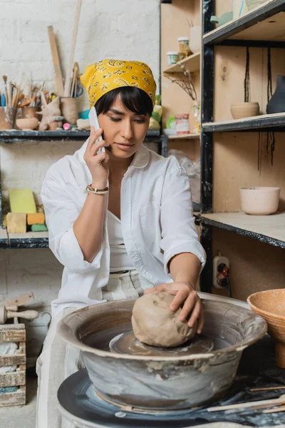 Young asian female artisan in headscarf and workwear talking on smartphone and putting clay on pottery wheel in ceramic workshop, artisan crafting ceramics in studio — Stock Photo