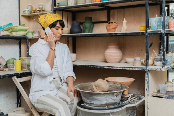 Young asian female potter in workwear and headscarf talking on smartphone while sitting near clay on pottery wheel and tools and working in pottery studio, artisan crafting ceramics in studio — Stock Photo