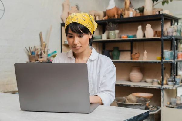 Young brunette asian female artist in headscarf using laptop at table while working in blurred ceramic workshop at background, pottery artist showcasing craft — Stock Photo