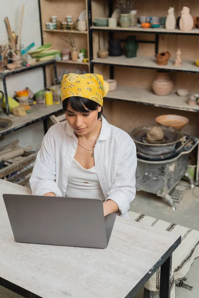 Vista de ángulo alto de la joven mujer asiática artesana en pañuelo para la cabeza y ropa de trabajo usando el ordenador portátil mientras está sentado en la mesa y trabajando en un taller de cerámica borrosa, artista de cerámica mostrando artesanía - foto de stock