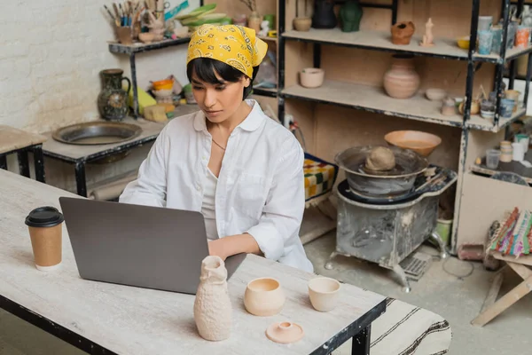Young brunette asian craftswoman in headscarf and workwear using laptop while working near coffee to go and clay products on table in ceramic workshop, pottery artist showcasing craft — Stock Photo