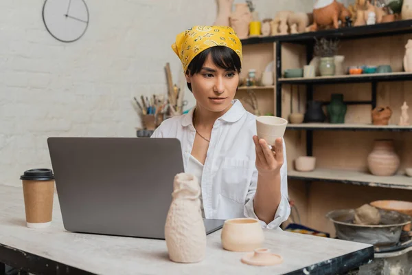 Jovem asiática morena artesã em workwear segurando produto de argila perto de laptop e café para ir à mesa em oficina de cerâmica borrada no fundo, artista de cerâmica apresentando artesanato — Fotografia de Stock