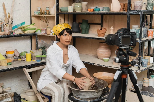 Jovem asiática feminina artesã em véu e workwear colocando argila na roda de cerâmica perto da câmera digital no tripé na oficina de cerâmica, artista de cerâmica apresentando artesanato, influenciador — Fotografia de Stock