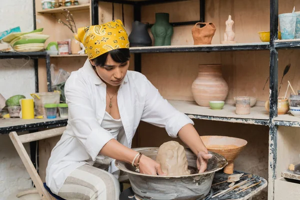 Jovem asiática artesã em vestuário de trabalho e lenço de cabeça moldando argila na roda de cerâmica perto de esculturas em rack e ferramentas em oficina de cerâmica, artista de cerâmica apresentando artesanato — Fotografia de Stock