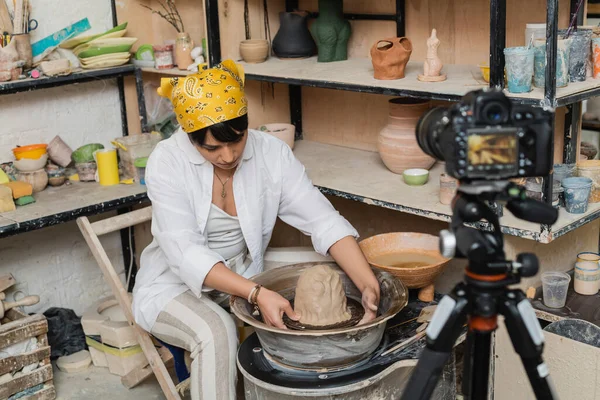 Young asian female artisan in workwear and headscarf molding clay on pottery wheel near blurred digital camera on tripod in ceramic workshop, pottery artist showcasing craft — Stock Photo