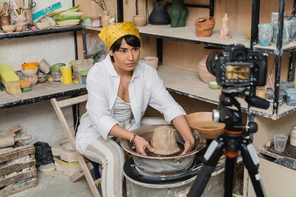 Young asian brunette craftswoman in headscarf and workwear molding clay on pottery wheel near digital camera and rack with sculptures at background, pottery artist showcasing craft — Stock Photo