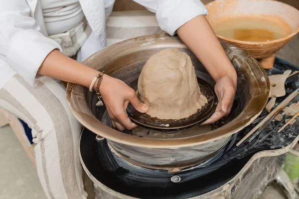 Cropped view of young female artisan in workwear working with clay on pottery wheel, tools and bowl with water at background in ceramic workshop, pottery artist showcasing craft — Stock Photo