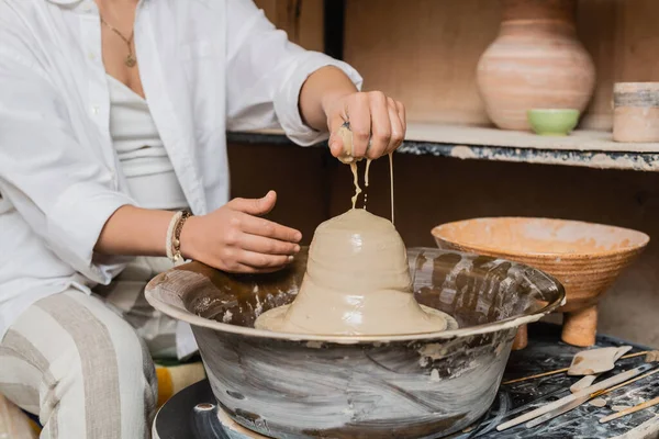 Cropped view of blurred female artist in workwear pouring water on clay on pottery wheel near tools and bowl in ceramic workshop at background, pottery artist showcasing craft — Stock Photo