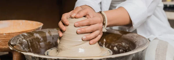 Cropped view of young female artisan in workwear molding wear clay while working on pottery wheel near bowl in ceramic workshop, pottery studio workspace and craft concept, banner — Stock Photo
