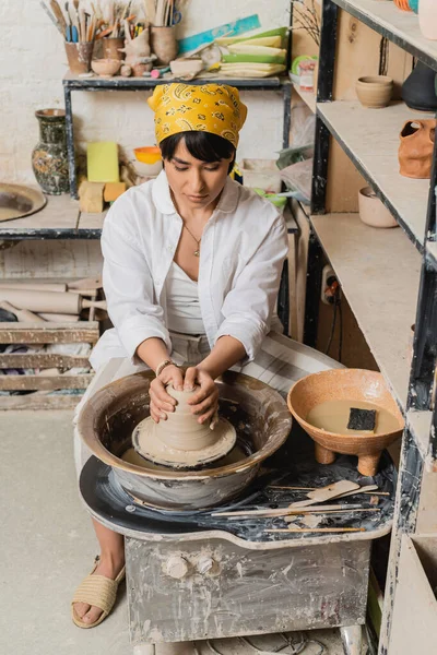 Young asian female artisan in headscarf in workwear molding clay on pottery wheel near bowl with water and sponge near rack in ceramic workshop, pottery studio workspace and craft concept — Stock Photo