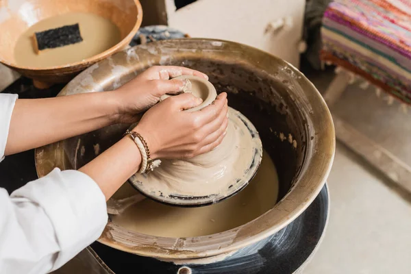 Ausgeschnittene Ansicht einer jungen Kunsthandwerkerin, die in Keramikwerkstatt, Töpferatelier und Bastelkonzept nassen Ton auf Töpferscheibe in der Nähe einer verschwommenen Schüssel mit Wasser und Schwamm formt — Stockfoto