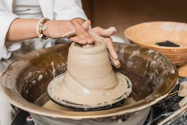 Cropped view of young female artist molding wet clay on pottery wheel while working near blurred bowl with water and tools in workshop, pottery studio workspace and craft concept — Stock Photo