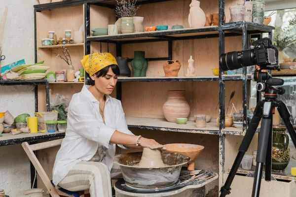 Jeune artisan asiatique en foulard moulant de l'argile humide sur une roue de poterie près d'un appareil photo numérique sur trépied dans un atelier de céramique, un atelier de poterie et un concept artisanal — Photo de stock
