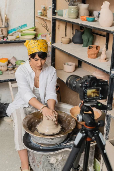 Young brunette asian artist in headscarf molding wet clay and working on pottery wheel near blurred digital camera on tripod in ceramic workshop, pottery studio workspace and craft concept — Stock Photo
