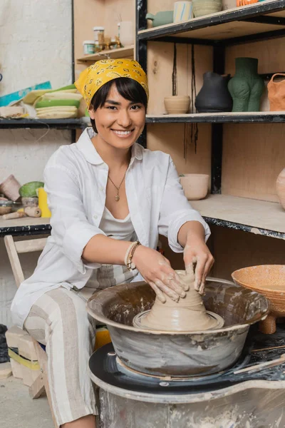 Веселая молодая азиатская художница в головном уборе, формирующая влажную глину, работая над гончарным колесом и глядя на камеру в керамической мастерской, керамической мастерской, рабочем пространстве и концепции ремесла — стоковое фото