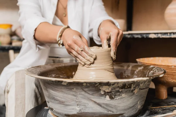 Cropped view of young female artisan in workwear shaping wet clay while working on pottery wheel near bowl at background, pottery studio workspace and craft concept — Stock Photo
