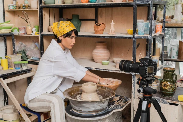 Asiatische Töpferin in Arbeitskleidung und Kopftuch blickt auf Digitalkamera und arbeitet mit Schale mit Wasser und nassem Ton auf Töpferscheibe, Töpferatelier und Bastelkonzept — Stockfoto