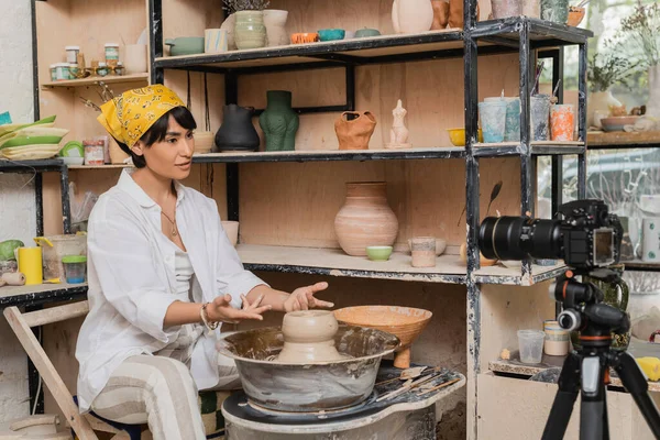 Asian female artisan in headscarf and workwear talking at digital camera on tripod while working with wet clay on pottery wheel near rack in art workshop, clay sculpting process concept — Stock Photo