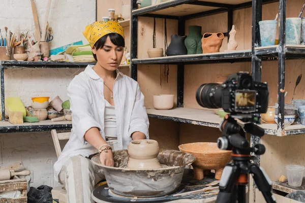 Young asian artist in headscarf and workwear working with wet clay on pottery wheel near blurred digital camera on tripod in ceramic workshop, clay sculpting process concept — Stock Photo