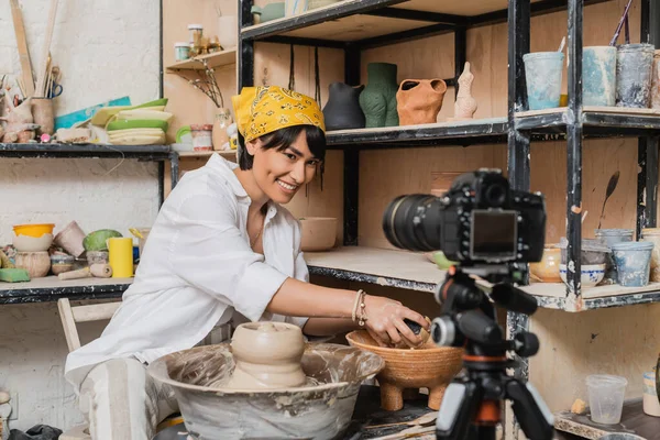 Улыбающаяся азиатская ремесленница в платке, держащая губку возле чаши с водой, глиной и гончарным колесом и глядя на цифровую камеру в художественной мастерской, концепция процесса глиняной скульптуры — стоковое фото