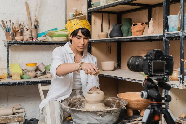 Junge asiatische Töpferin mit Kopftuch gießt Wasser aus Schwamm auf Ton auf Töpferscheibe neben verschwommener Digitalkamera in Keramik-Werkstatt, Tonbildhauerei-Konzept — Stockfoto