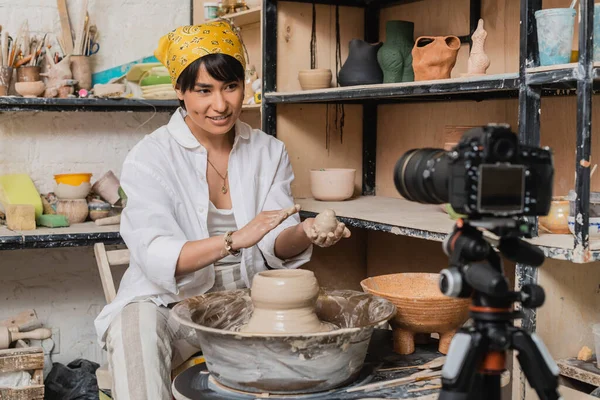 Positive asian female artist in headscarf holding wet clay and looking at digital camera on tripod near pottery wheel in art workshop, clay sculpting process concept — Stock Photo