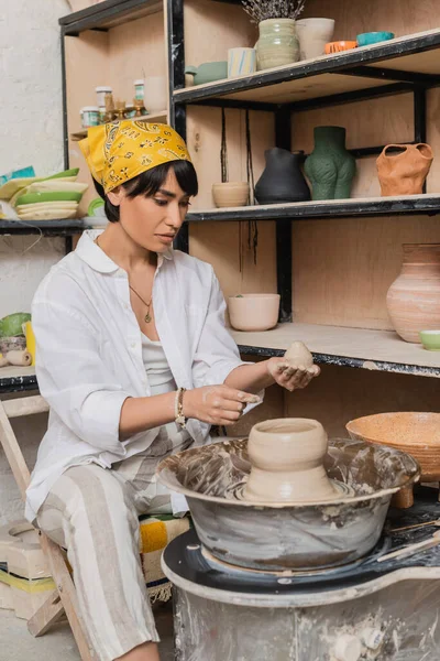 Young asian female artisan in headscarf holding clay near pottery wheel and rack with sculptures in ceramic workshop at background, clay sculpting process concept — Stock Photo