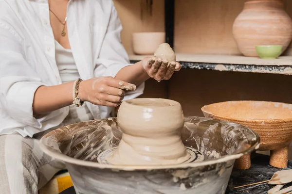 Ausgeschnittene Ansicht einer jungen Kunsthandwerkerin in Arbeitskleidung, die Ton auf Töpferscheibe hält und gießt und in der Nähe einer Schüssel in einer Keramikwerkstatt arbeitet, Konzept zur Tonbildhauerei — Stockfoto