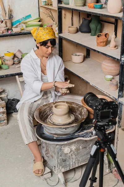 Young asian artisan in headscarf and workwear holding wet clay and working near pottery wheel and digital camera on tripod in ceramic studio, clay sculpting process concept — Stock Photo