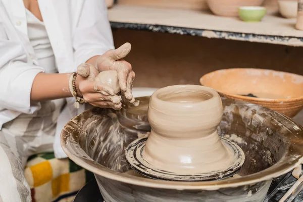Cropped view of young female potter in workwear holding wet clay near pottery wheel and blurred bowl in blurred ceramic studio, clay sculpting process concept — Stock Photo