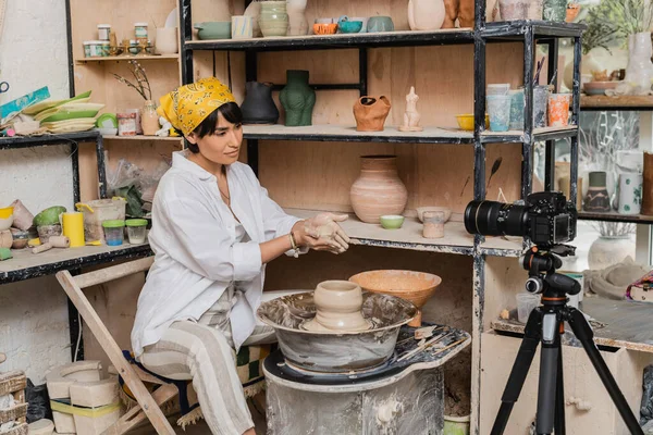 Young asian female artisan in workwear and headscarf holding wet clay near digital camera on tripod and pottery wheel in ceramic workshop, clay sculpting process concept — Stock Photo