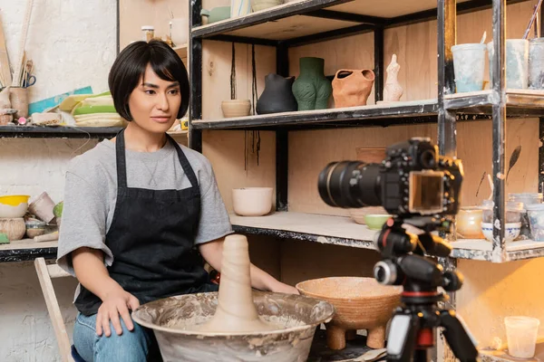Young asian brunette craftswoman in apron looking at blurred digital camera on tripod near wet clay on pottery wheel and rack in ceramic workshop, pottery tools and equipment — Stock Photo