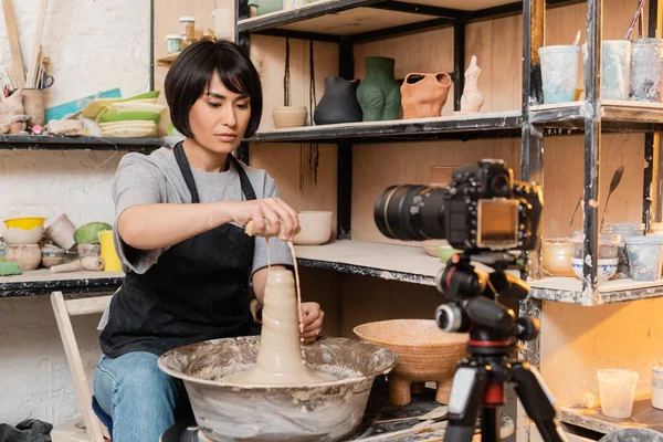 Young brunette asian female artisan in apron pouring water from sponge on wet clay and pottery wheel near digital camera on tripod in studio at background, pottery tools and equipment — Stock Photo