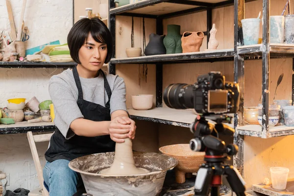 Young asian brunette craftswoman in apron molding wet clay on pottery wheel near blurred digital camera and sculptures on rack in workshop, pottery tools and equipment — Stock Photo