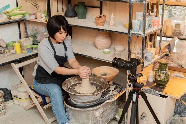 Молодая азиатская женщина ремесленница в фартуке формирования влажной глины на гончарном круге возле чаши с водой и инструменты рядом с цифровой камерой в керамической мастерской на заднем плане, керамические инструменты и оборудование — стоковое фото